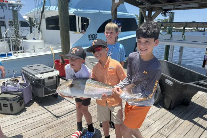 Four boys on a dock holding a large fish, with boats in the background.