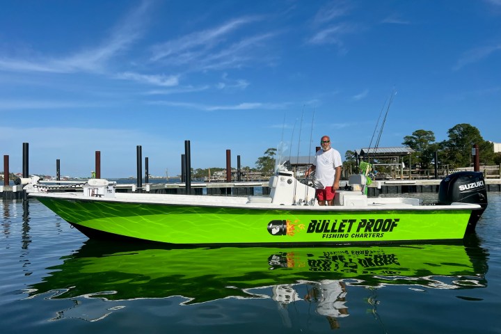 Man standing on a bright green fishing boat named 'Bullet Proof' in a marina.