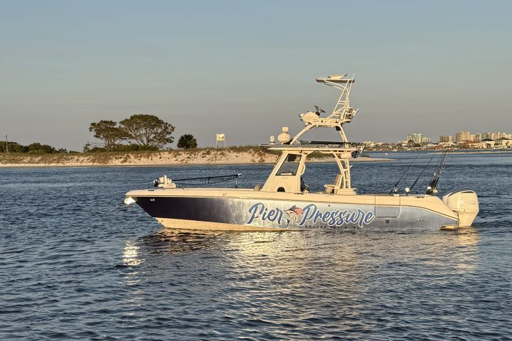 Boat named 'Pier Pressure' on water near shoreline with distant trees and buildings.