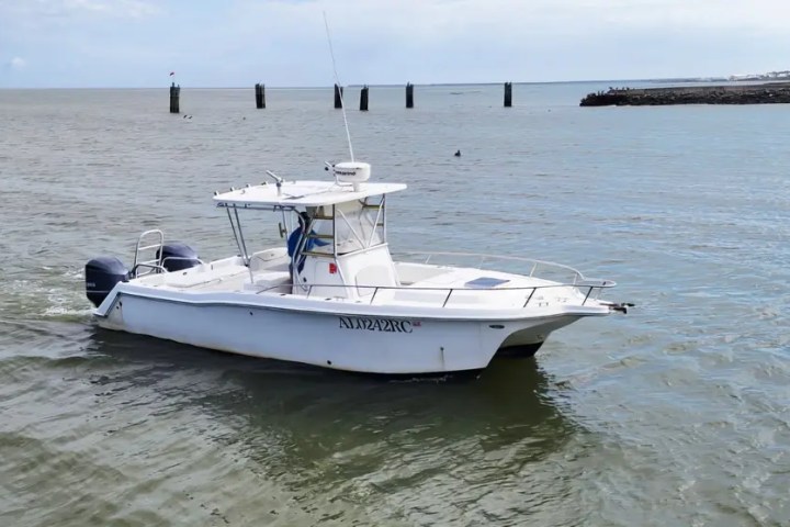 A white motorboat with twin engines on a calm sea, near pilings and a distant shoreline.
