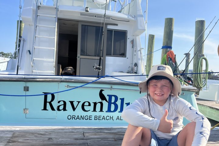 Child in a hat sitting on a dock next to a boat named RavenB.